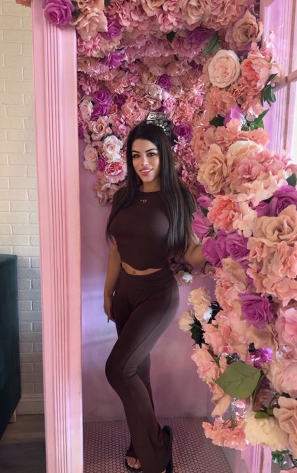 A young woman with long dark hair wearing a brown two-piece outfit and a tiara, posing in front of a pink floral wall in what appears to be a photo booth.