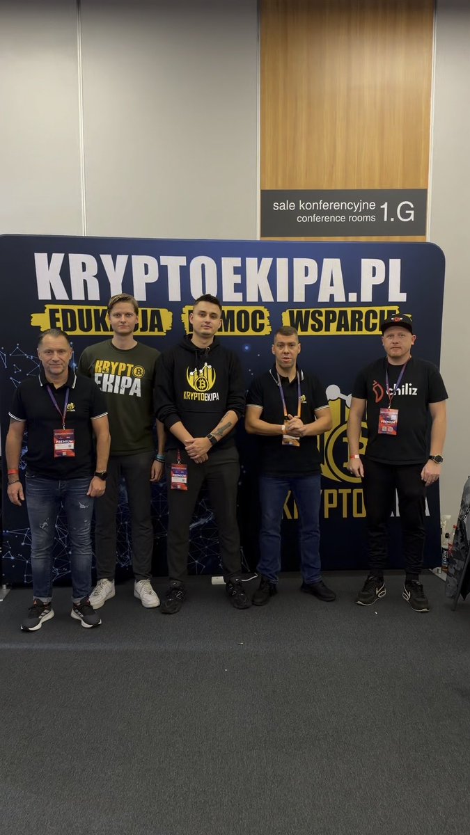 Five men pose in front of a 'KRYPTOEKIPA.PL' banner with the words 'EDUKACJA POMOC WSPARCIE' visible, near a sign indicating 'sale konferencyjne conference rooms 1.G'.