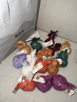 Photo of numerous colorful fabric bundles (purple, green, orange, brown) with tags tied to them, scattered on a tiled floor next to a gray bag.