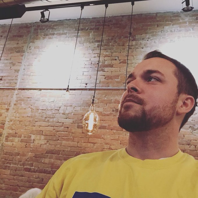 A bearded young man in a yellow shirt looking upward in a room featuring exposed brick walls and hanging Edison-style light bulbs.