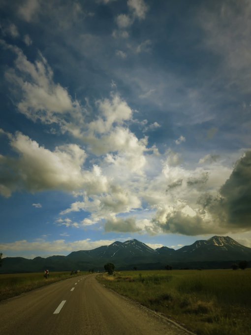 The image shows a road with white lines down its center, surrounded by grassy fields and mountains in the background. There are no people visible in the photo. The sky above has clouds, suggesting that it might be a cloudy day or during a transitional phase of weather conditions. The text "GM future millionaires Green everywhere Are you guys ready for new highs" is related to the post's title, which could imply that the person who posted this image is an optimist and wants to share their enthusiasm about the potential benefits of investing in green energy or sustainable living.