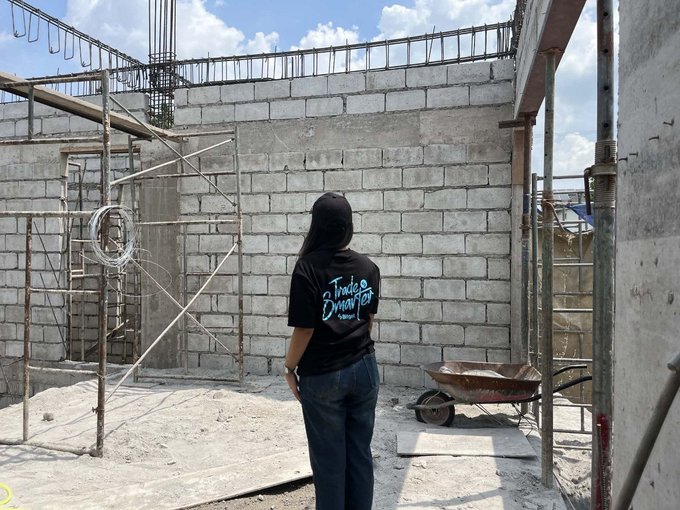 A person with long dark hair, wearing a black t-shirt with blue text reading 'Trade is smart for...' and blue jeans, stands with their back to the camera at a concrete construction site with rebar walls and a wheelbarrow.