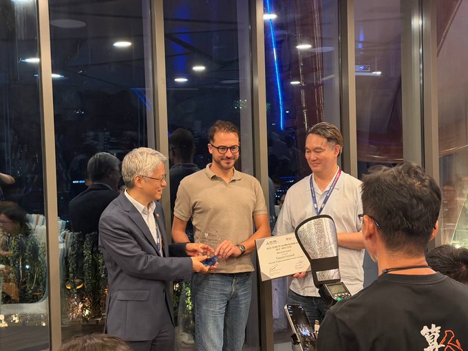 People exchange a plaque and a trophy-like award at an indoor ceremony with large windows in the background.