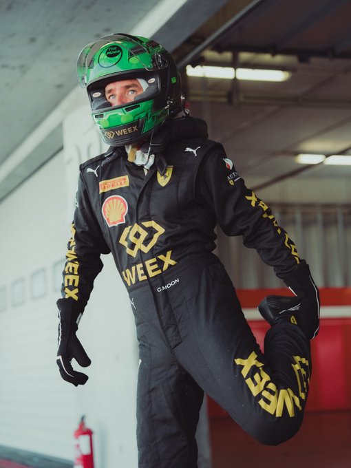 A race car driver wearing a black racing suit with Puma, Shell, and WEEX sponsor logos, posing in a garage while wearing a green helmet with Monster Energy branding.