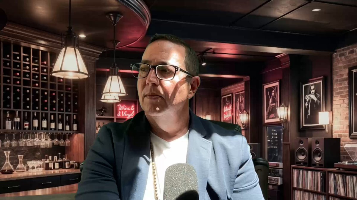 The image features a man wearing glasses, standing in front of a bar with wine bottles displayed behind him. He appears to be talking into a microphone or giving a speech at an event. The background includes a brick wall and several lights hanging from the ceiling, creating a cozy atmosphere for the gathering. There is also a sign that reads "Ripple/CEO Gives XRP FORMULA youtu.be/JSdjVLROubg".