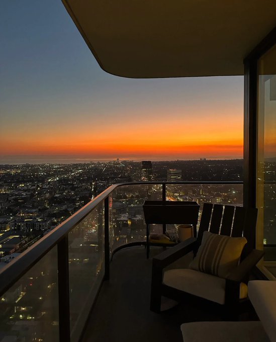 A view from a high-rise balcony at sunset, showing a cityscape with twinkling lights below and two chairs in the foreground.