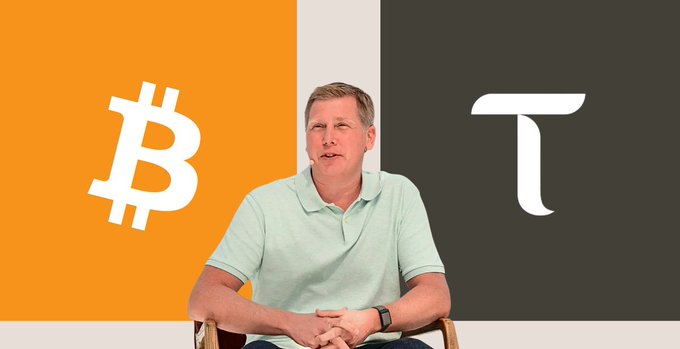 A man in a light green polo shirt sits in front of a backdrop displaying the Bitcoin logo (orange side) and Pi logo (black side).