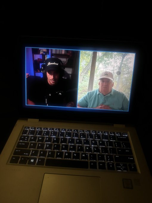 A laptop in a dark room displaying a video call between two men: a younger man wearing a black 'Pinehurst' cap on the left, and an older man in a green polo and white cap on the right.