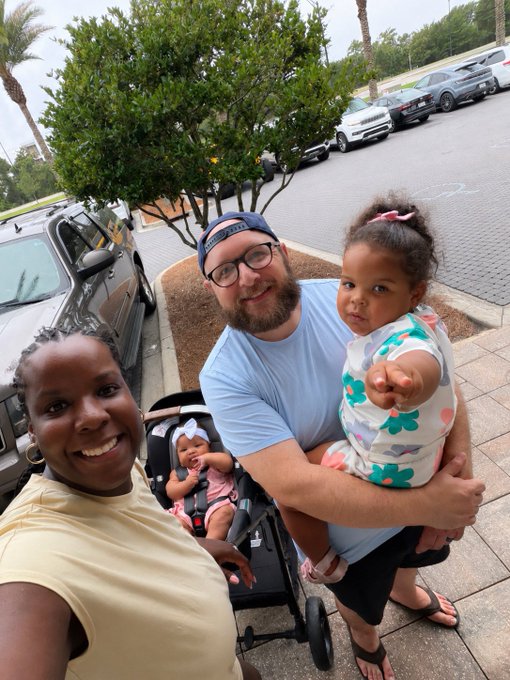 A smiling family of four takes a selfie in a parking lot, with dad holding their young daughter and another baby asleep in a stroller. The joyful family photo reflects the personal, family-oriented side of the poster's introduction as a 34-year-old graphic designer and coffee-loving NFT enthusiast.