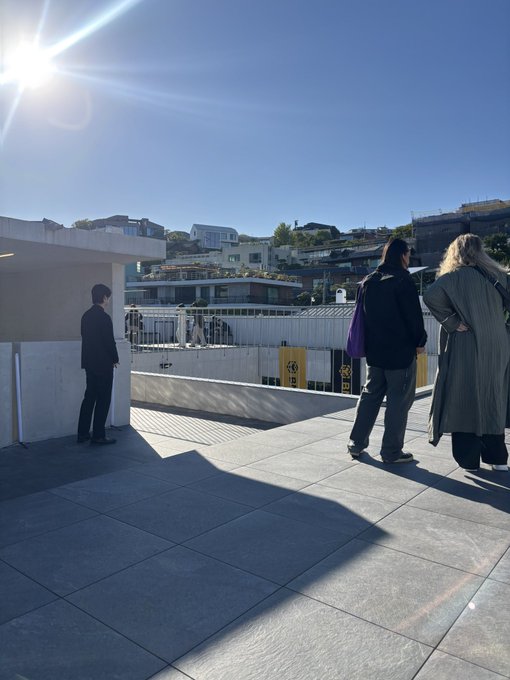 Three people stand on a sunlit rooftop terrace overlooking a hillside neighborhood with the sun flaring brightly in the upper left corner.