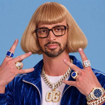 A person with a blonde bob wig, glasses, and a blue velour jacket displays hands covered in large rings and gold chains with a 'OG' pendant.