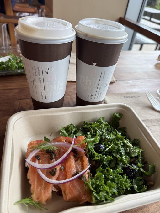 Two Philz coffee cups sit on a table behind a takeout container holding smoked salmon topped with red onions and capers alongside a kale and blueberry salad.