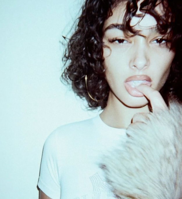 A fashion-style photograph of a young woman with short curly dark hair and a white t-shirt, touching her finger to her mouth with a slight expression, wearing gold hoop earrings.
