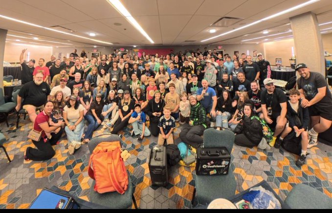 A large group photo captures dozens of attendees, including children, gathered together in a conference hall with patterned carpet, posing behind several pieces of luggage.