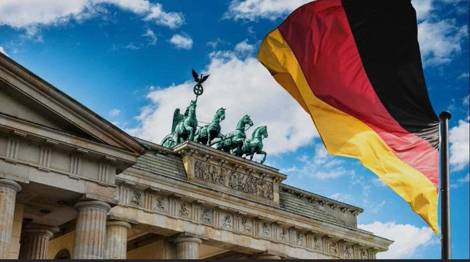 The Brandenburg Gate in Berlin is shown beneath a blue cloudy sky, with the green Quadriga statue visible on top and the German flag waving prominently in the foreground.