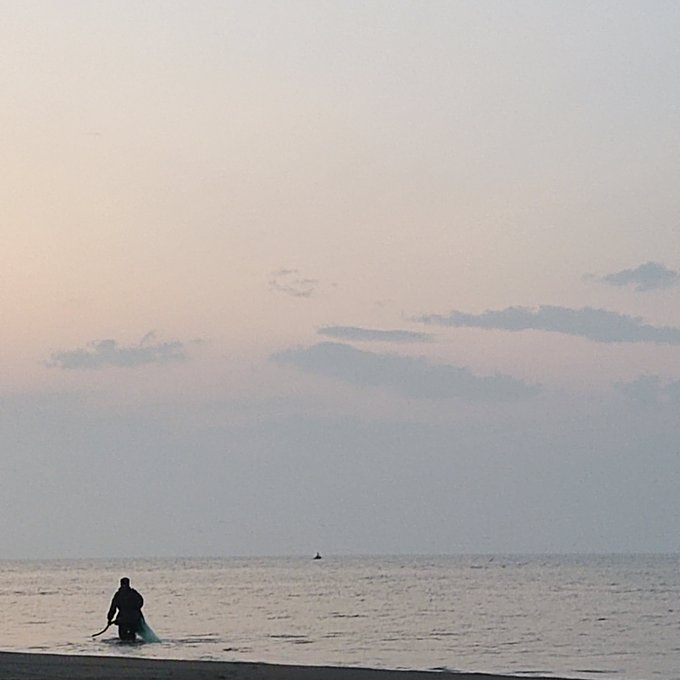 A silhouetted figure wades through the calm ocean at dusk, carrying what appears to be a surfboard or fishing net, under a soft pastel sky with faint clouds. The serene, contemplative scene evokes the quiet moment that the post's caption "G.m" (good morning) suggests—an early, peaceful start to the day by the sea.