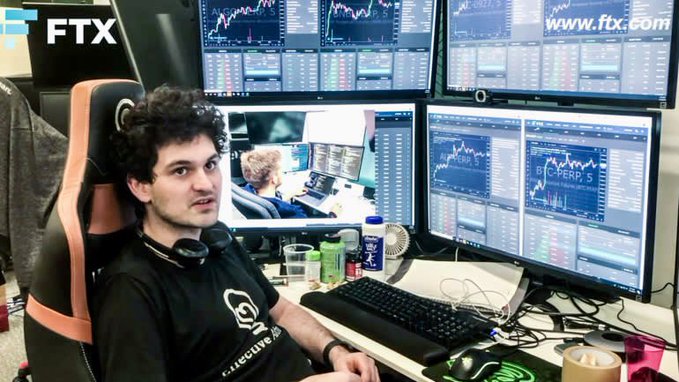 FTX founder Sam Bankman-Fried sitting in front of multiple trading monitors at an FTX trading desk.