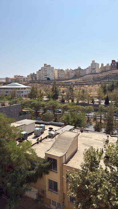 A daytime cityscape view showing residential buildings on a hillside, with green trees, a road, and rooftops in the foreground.