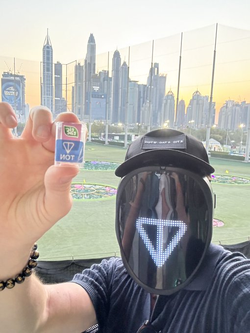 A person wearing an LED face mask and 'HOPE GATE OFFZ' cap holds up a 'TOP' sticker, standing before a green field with a city skyline in the background.