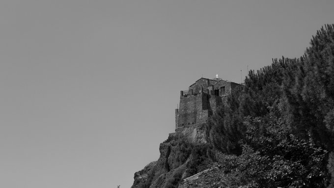 A black and white photo of a stone building or castle perched on a steep, rocky hillside covered with vegetation.