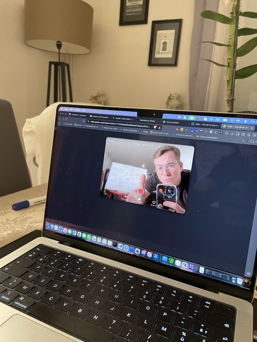 A MacBook laptop on a desk displays a video recording of a man wearing glasses holding up a handwritten document with equations and graphs, with a recording timer visible.