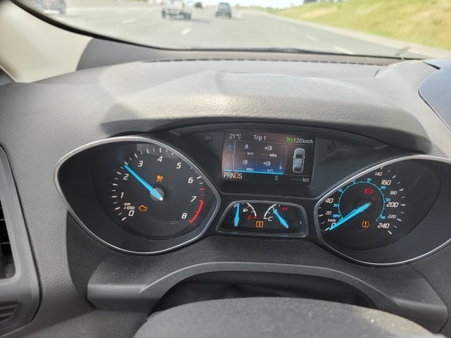 The dashboard shows a car in motion with several illuminated warning lights, including the check engine light, traction control, low tire pressure (PMS), and an open door indicator, with the speedometer and tachometer needles actively registering movement.