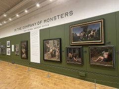 An art gallery wall displays several framed paintings under the heading 'IN THE COMPANY OF MONSTERS' in a well-lit exhibition space.