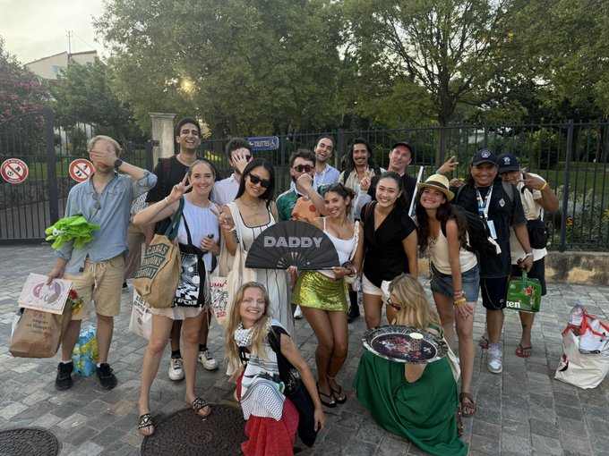 A large group of young adults poses outdoors on a city street, with one woman in the center holding a fan that reads 'DADDY'.
