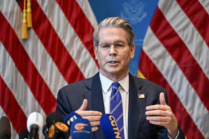 U.S. Secretary of the Treasury Scott Bessent speaks at a press conference, gesturing with his hands while standing at a podium with microphones from various outlets (including FES and CGTN), with American flags and the Treasury seal behind him.