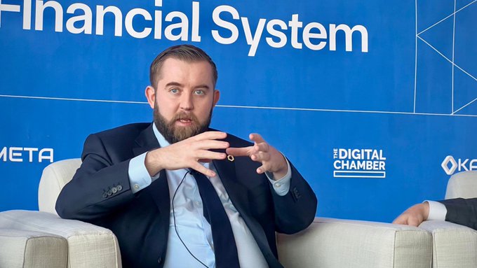 A bearded man in a dark suit speaks while gesturing with both hands at 'THE DIGITAL CHAMBER' event, with 'Financial System' visible on the blue backdrop behind him.