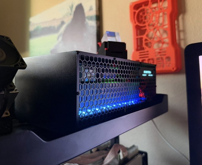 A black computer component, likely a power supply or small PC case, sits on a shelf with a honeycomb mesh front panel illuminated by blue LED lights, with an orange 3D-printed object mounted on the wall behind it.