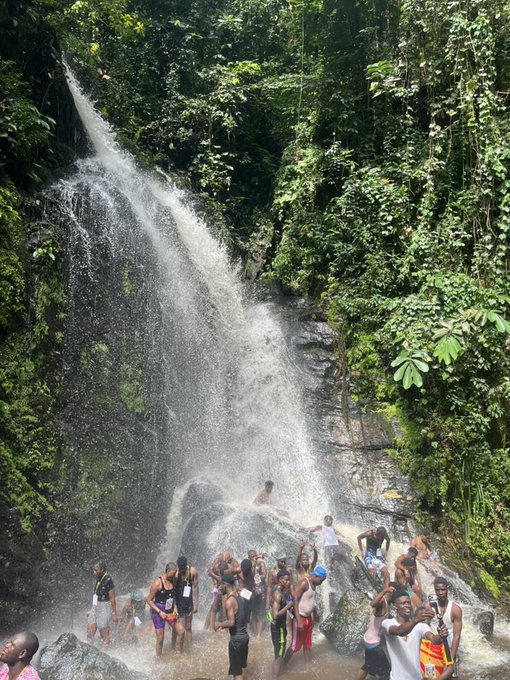 A group of people are gathered at the base of a waterfall in a lush green tropical setting, enjoying the water on a sunny day.
