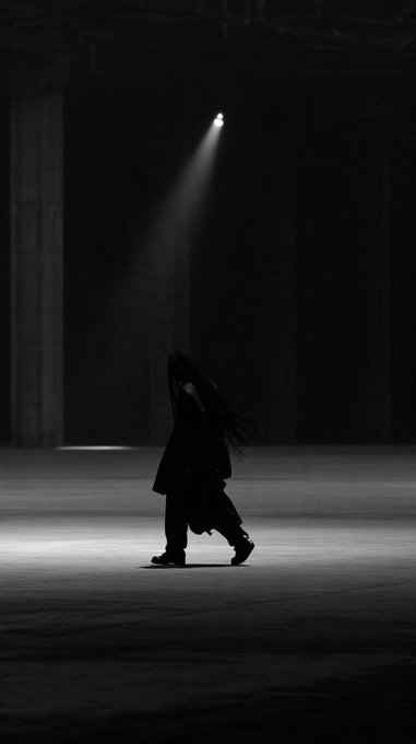 A black and white photograph shows a silhouetted figure with long hair walking across a large, dark industrial space illuminated by a single dramatic spotlight from above.