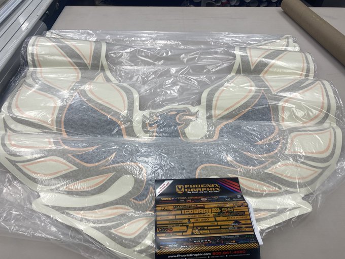A packaged motorcycle fairing in plastic wrap featuring 'Phoenix Graphics' branding and racing decals including 'Roberts', 'Hinson', and 'Suzuki'.