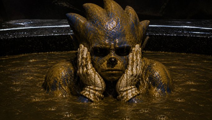 A detailed sculpture or model of a creature with spiky head features emerging from a pool of brown, mud-like liquid, with its hands pressed against its face.