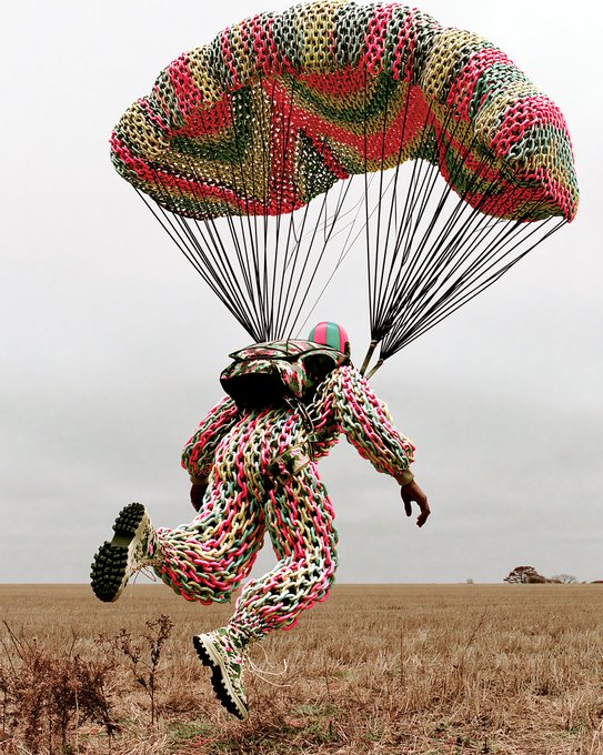 A surreal photograph of a person in a colorful knit outfit and helmet parachuting with a matching multi-colored parachute over a dry grassy field.