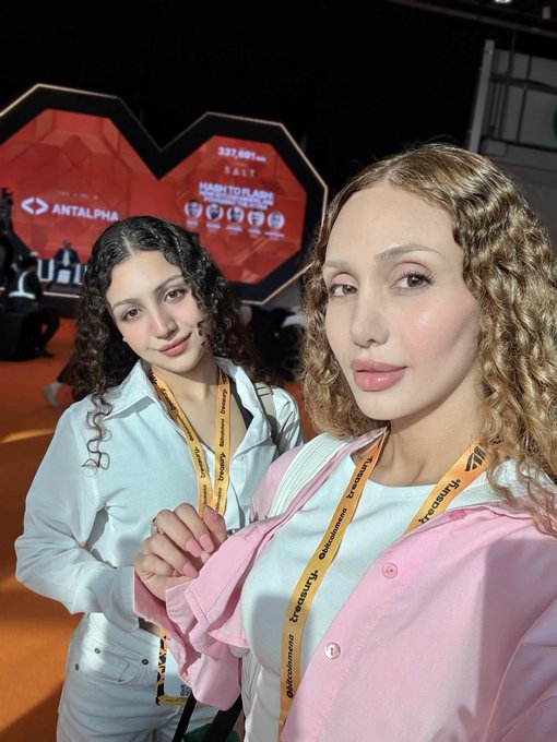 Two young women wearing "decubate" lanyards pose for a selfie at the TOKEN2049 crypto conference, with a stage backdrop displaying "237,691m SALT" and "ANTALPHA" branding visible behind them. The image captures them packing their bags for the event, fitting the post about networking with crypto projects and creators for potential collaborations.