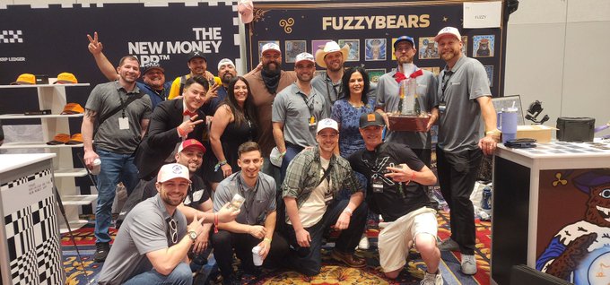 A large group of people pose happily together in a trade show booth setting, with signs reading "THE NEW MONEY" and "FUZZYBEARS" visible behind them.