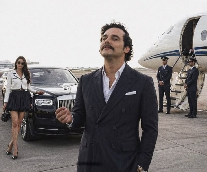 A man with a mustache in a dark double-breasted suit stands on a tarmac next to a black Rolls-Royce, with a woman in a white jacket and black skirt behind him, and pilots by a private jet in the background.