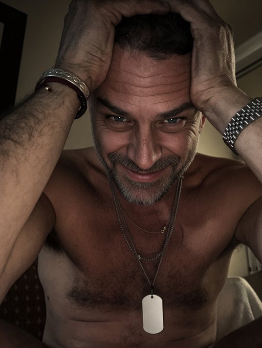 A selfie of a shirtless, muscular older man with grey hair and a goatee, holding his head with both hands and smiling at the camera, wearing dog tags and wristwatches.