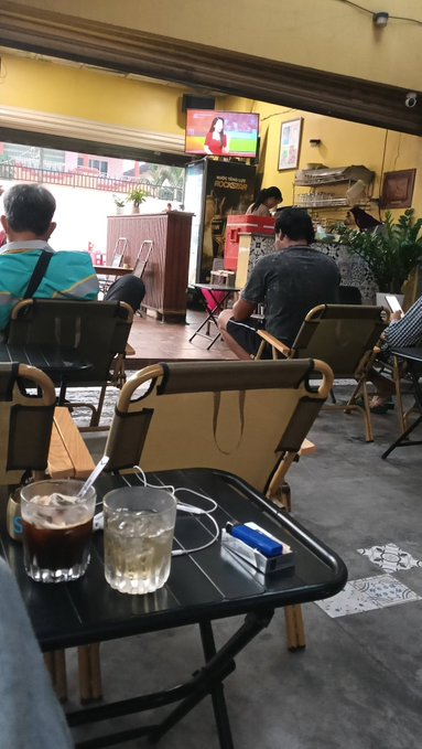 A casual café scene shows patrons watching a soccer match on TV, with two glasses—one of iced coffee and one of water—resting on a small table alongside a pack of cigarettes, capturing a relaxed morning moment that aligns with the post's "gm" and World Cup wake-up vibe.