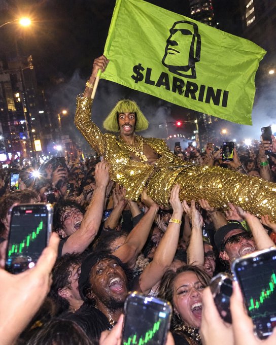 A person in a gold sequin dress and green wig is crowd-surfing while holding a green flag that reads '$LARRINI' with a Moai face logo, as fans hold up phones displaying rising stock charts.