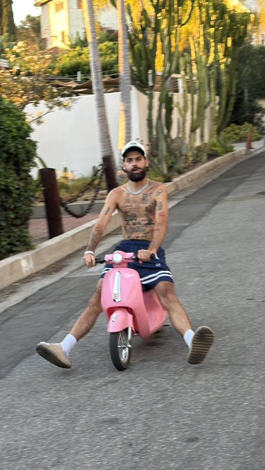 A shirtless, tattooed man wearing a cap and blue shorts does a split on a pink Vespa-style scooter down a palm-lined road, channeling the breezy, sun-soaked vibes of "Solana Summer."