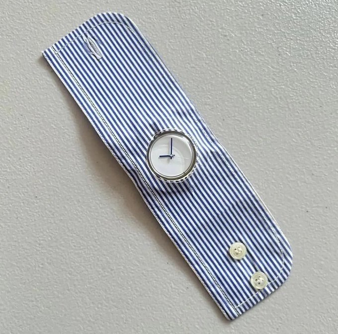 A wristwatch with a white face and silver casing is mounted on a blue and white striped fabric cuff featuring two button closures. The watch is displayed against a textured white background.