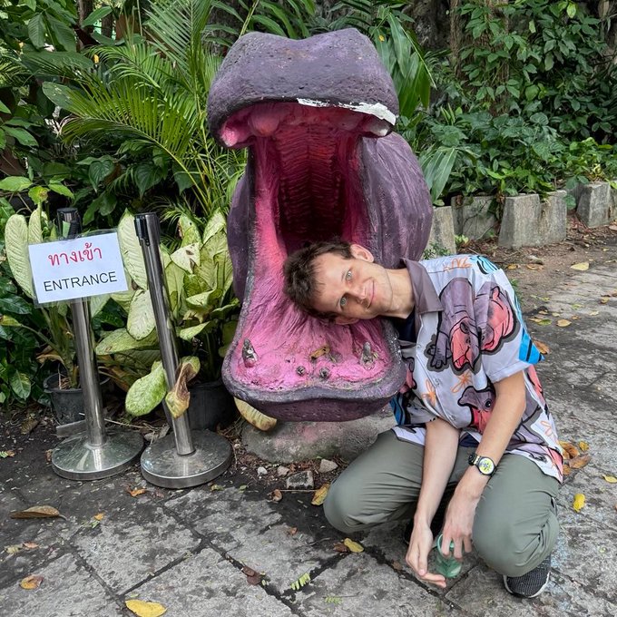 A man poses playfully next to the gaping "mouth" of a large purple and pink monster sculpture, with a sign in the foreground reading "ทางเข้า / ENTRANCE" in Thai and English. The post celebrates Vitalik Buterin's 11th anniversary with Ethereum, humorously framing the monster's open jaws as a fitting symbol for the wild journey of crypto's evolution.