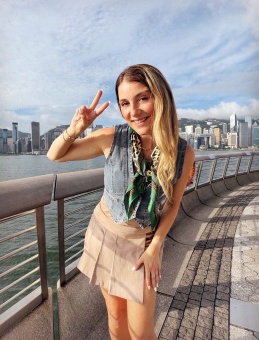 A young woman smiling and making a peace sign while posing on a waterfront promenade, with a city skyline and harbor visible in the background under a partly cloudy sky.