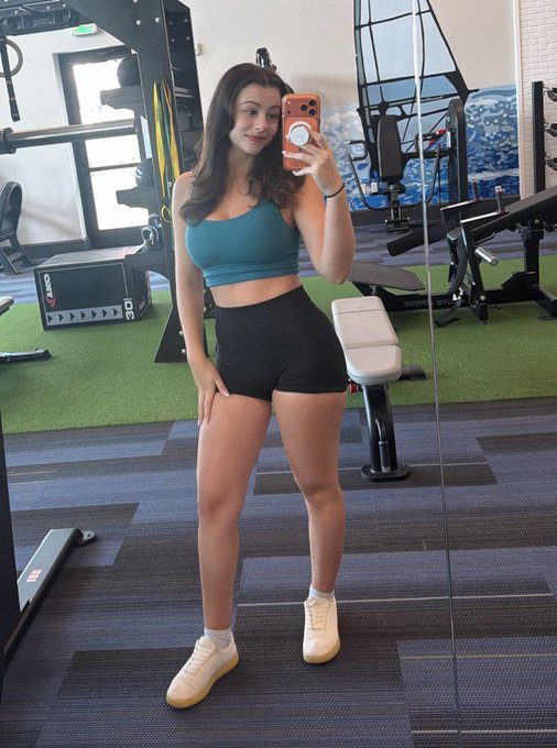 A woman takes a mirror selfie in a gym, wearing a teal sports bra, black shorts, and light-colored sneakers.