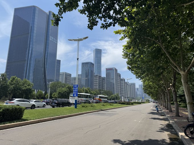 A street-level view of a city with modern high-rise buildings, trees lining the road, and a clear blue sky with some clouds.
