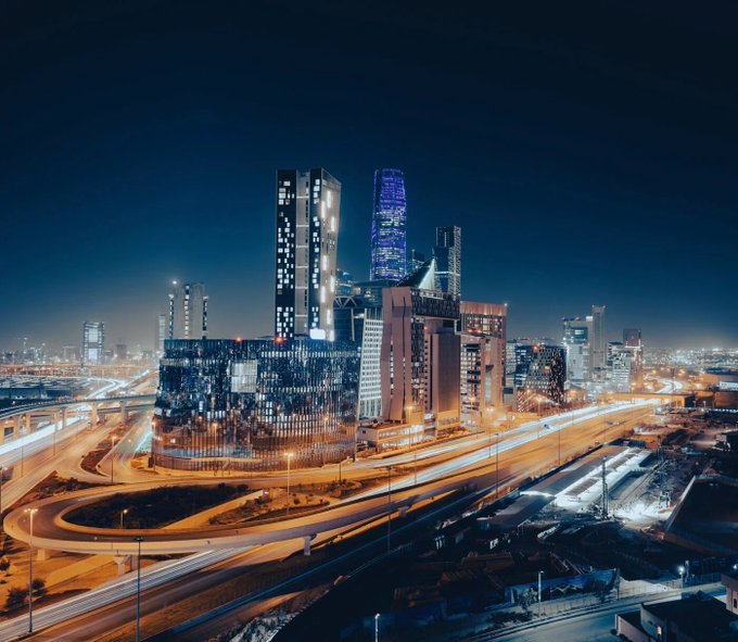 A nighttime cityscape showing illuminated skyscrapers and highways with light trails, featuring a tall building displaying 'OMAN' in lit windows.