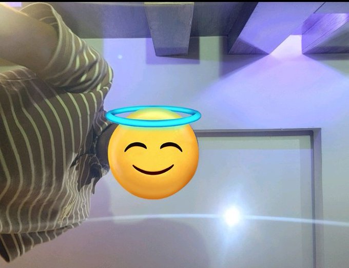 A low-angle selfie of a person wearing a striped shirt, with a smiling angel emoji face sticker digitally placed over their head, standing under a ceiling with exposed beams.